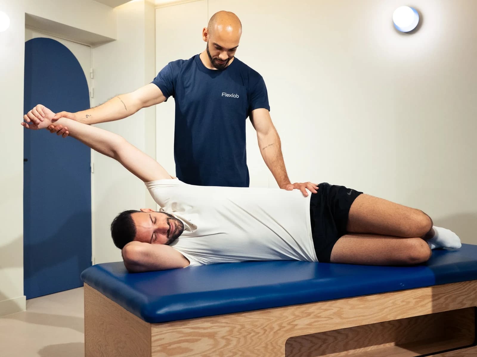 Un flexologue ouvre l’épaule d’un client allongé sur la table bleue, le bras tendu au-dessus de la tête.